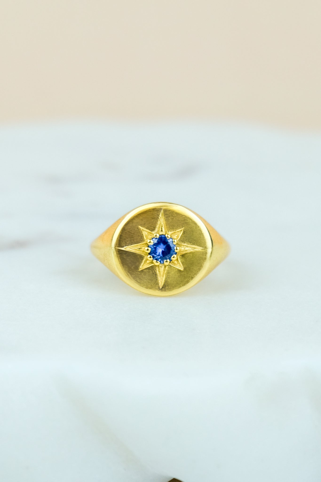 Gold Wayfinder signet ring with a blue gemstone and star engraving on a white marble background
