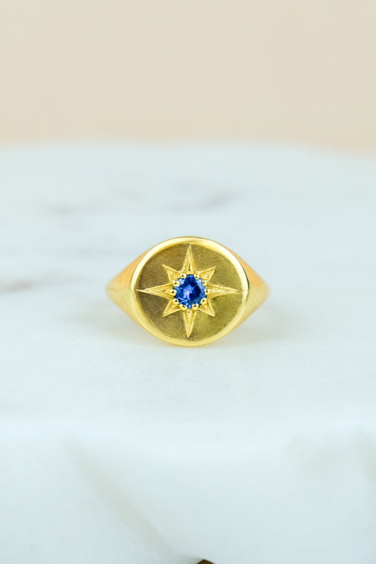 Gold Wayfinder signet ring with a blue gemstone and star engraving on a white marble background