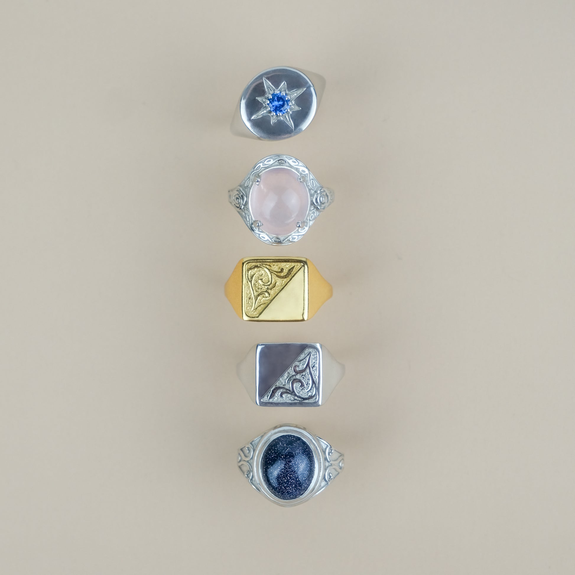 Five different signet rings with various designs and gemstones on a beige background