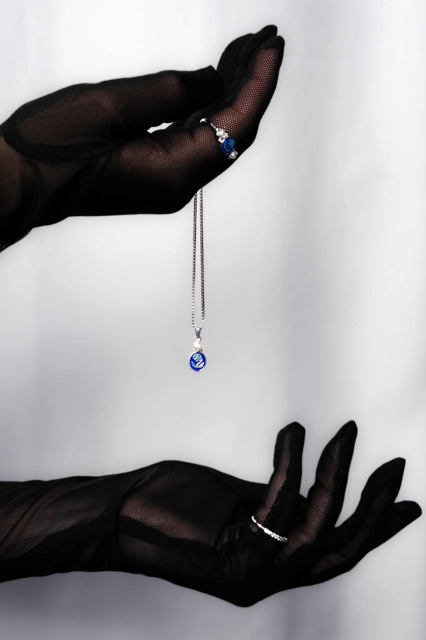 Black gloved hand dramatically holding Mourning Bloom sterling silver pendant, blue enamel forget-me-not necklace, Ether Jewelry