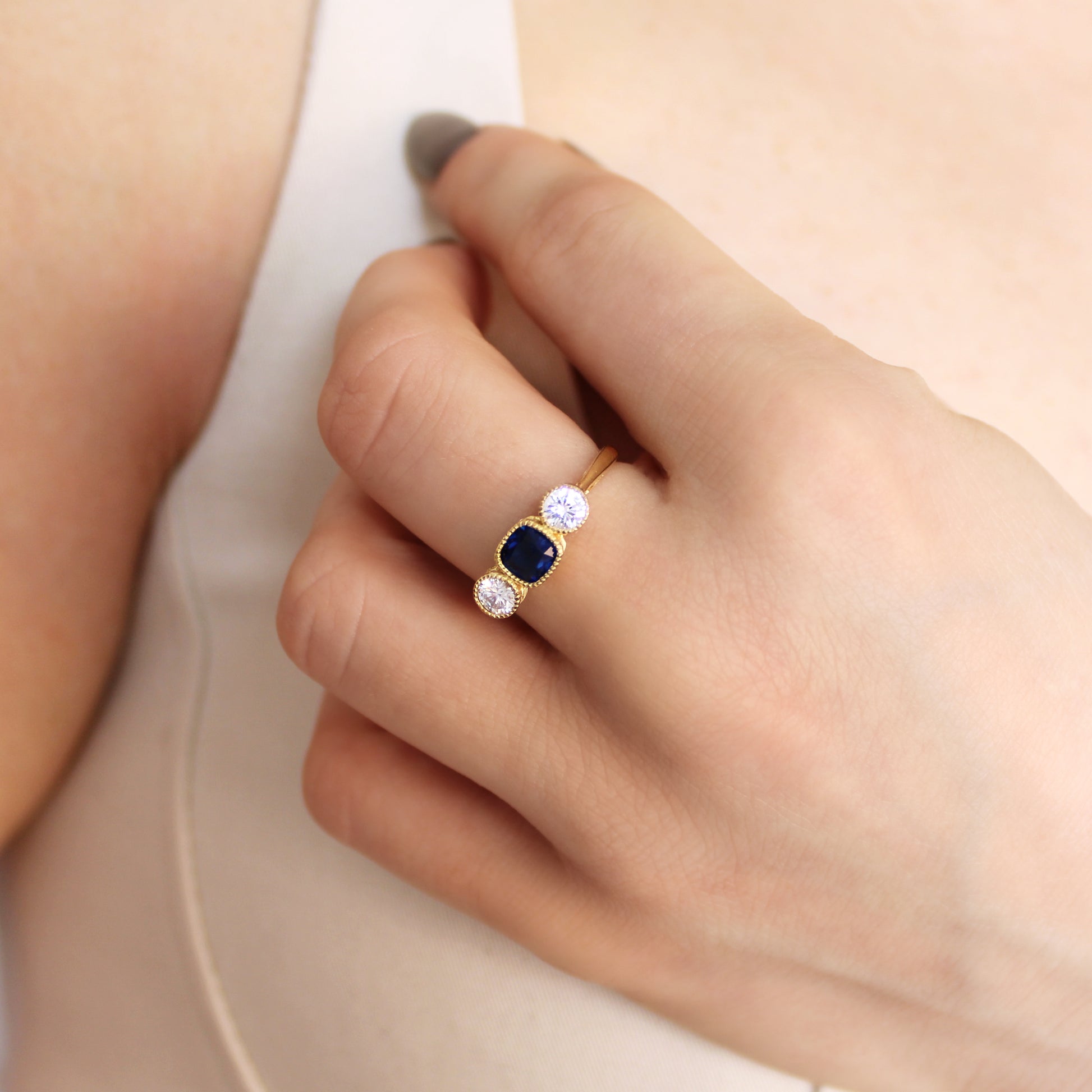 Edwardian Empress ring in 18k gold vermeil worn on model in beige dress, hand near chest, blue CZ three stone ring, Ether Jewelry