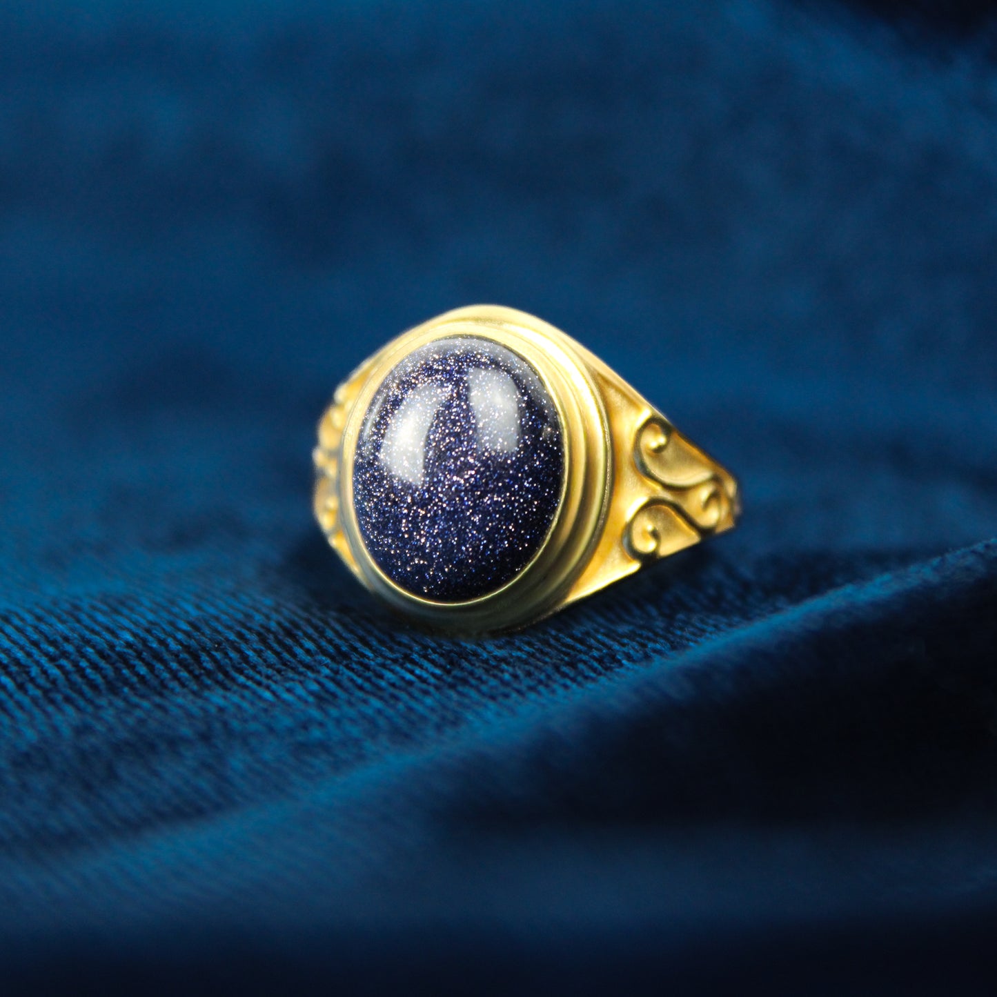 Gold night keeper signet ring with a deep blue sandstone gemstone on a blue fabric background
