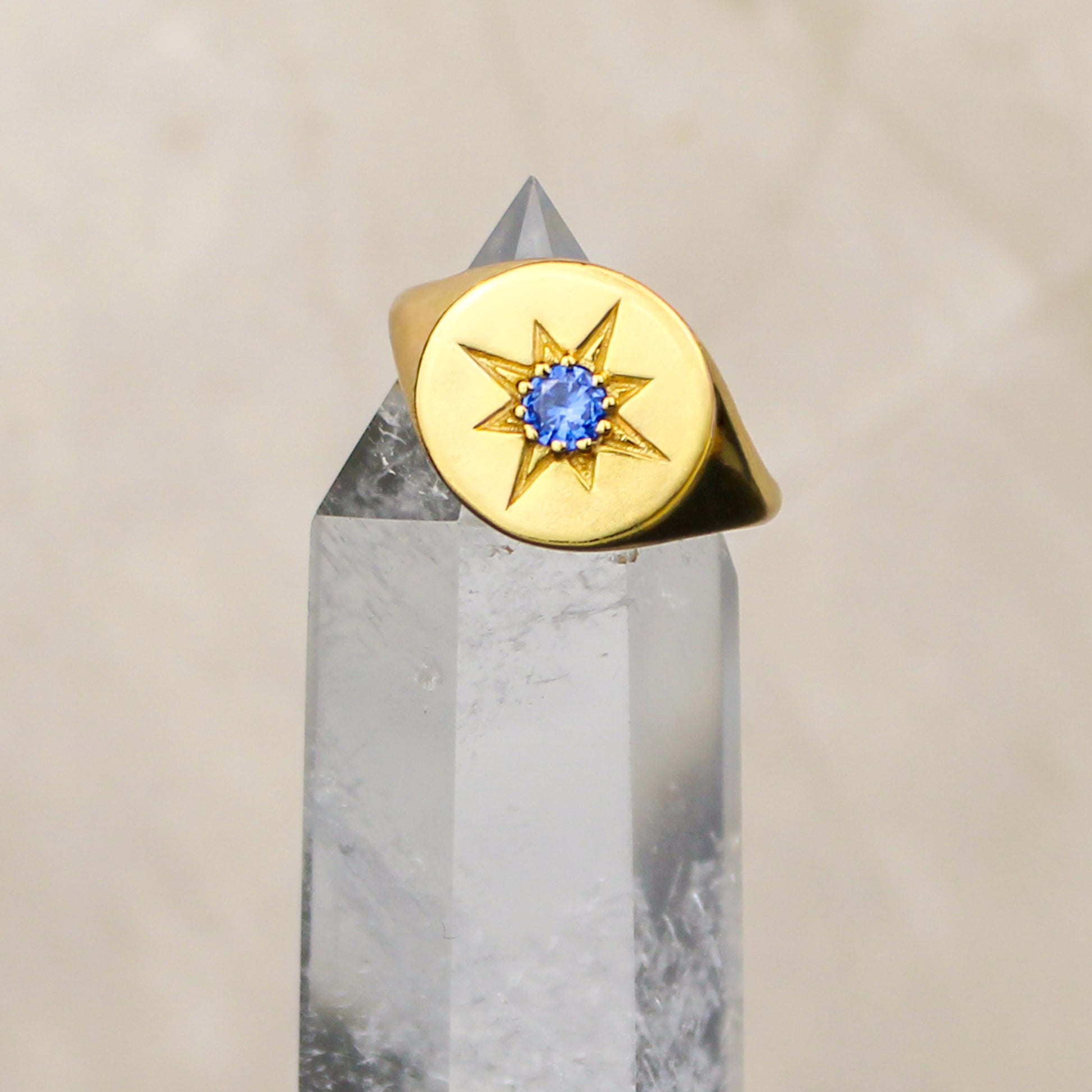 Gold Wayfinder signet ring with a blue gemstone and star engraving on a gray crystal prism against a beige background