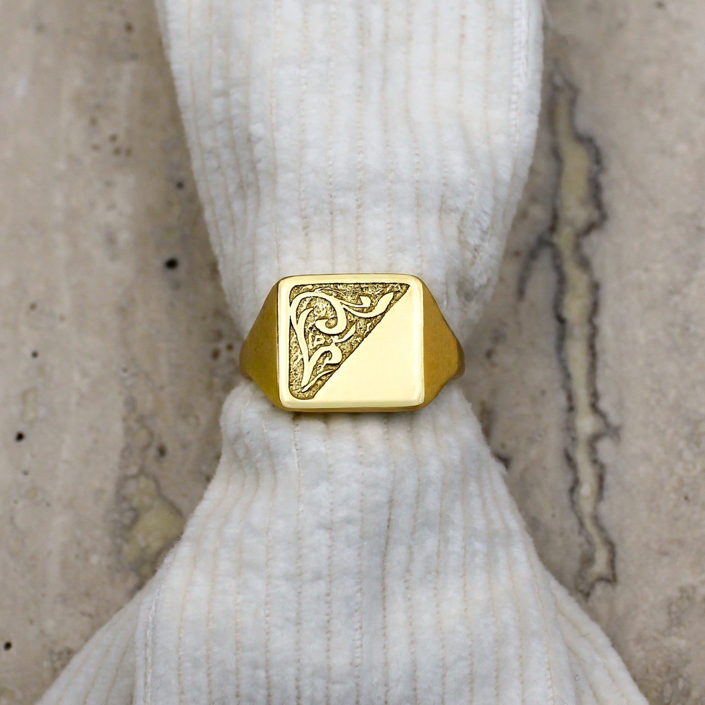 Gold square signet architect ring with intricate design on a white knitted fabric and marble background