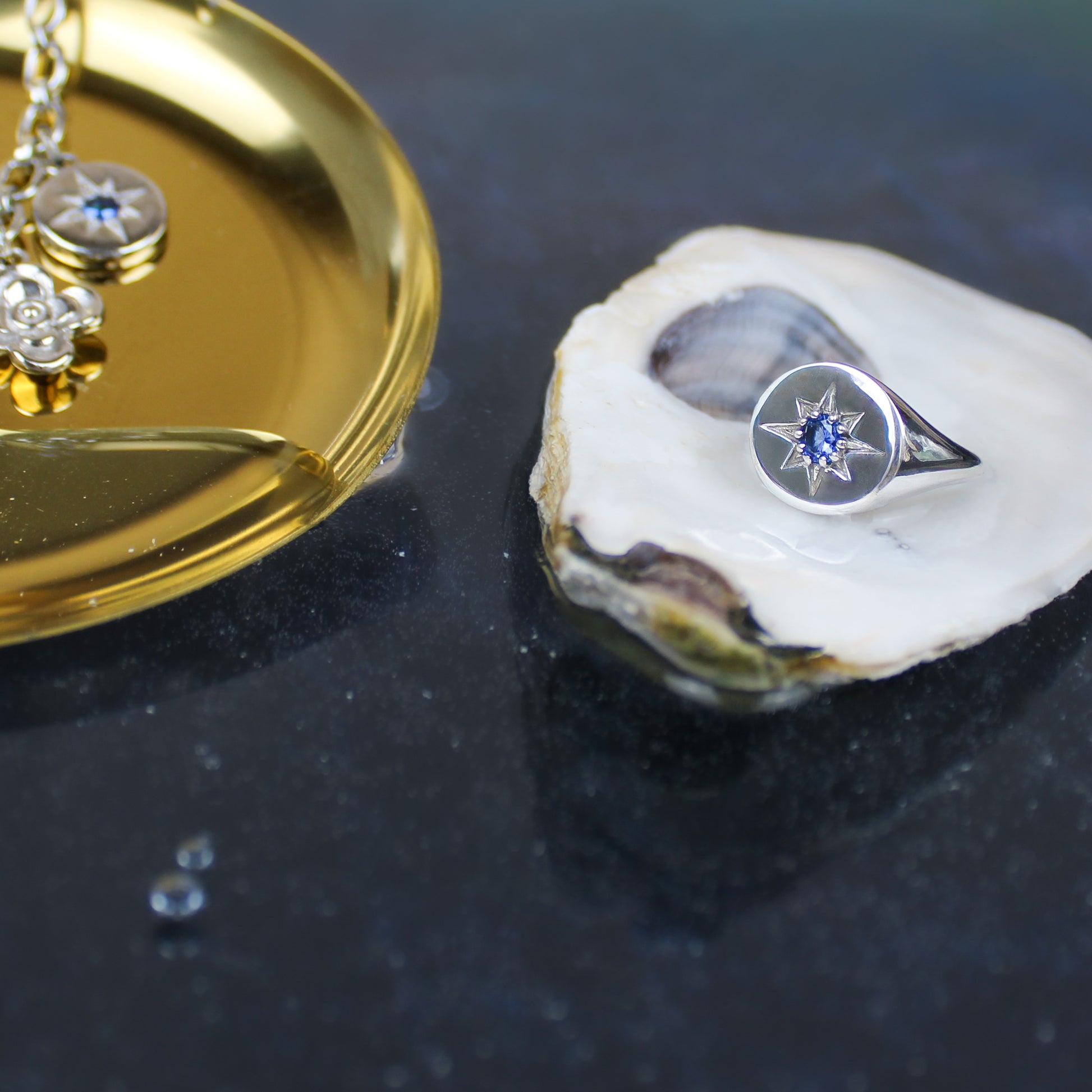 Sterling Silver signet ring with blue gemstone and star on a white shell next to a gold dish with jewelry