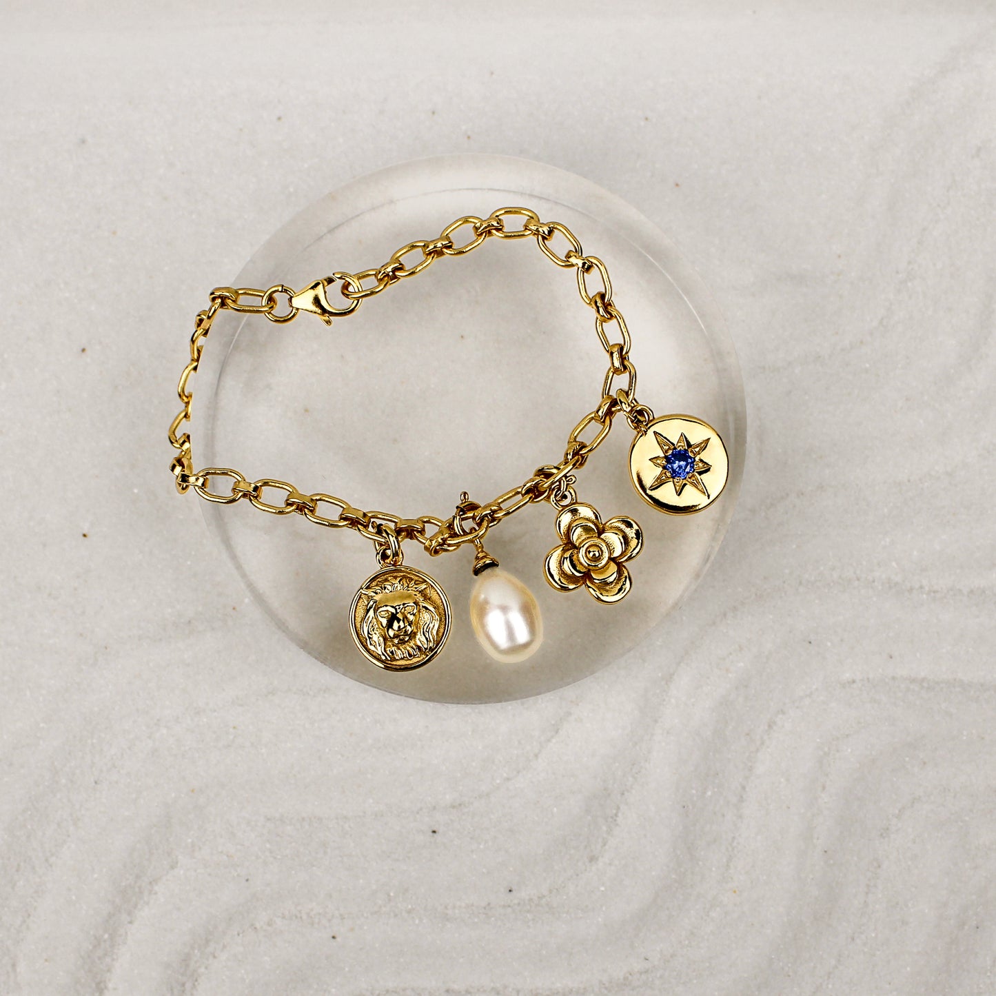 Archivist Bracelet 18k gold vermeil on clear dish over sand, multi-charm bracelet with lion, pearl, flower and north star charms, Ether Jewelry