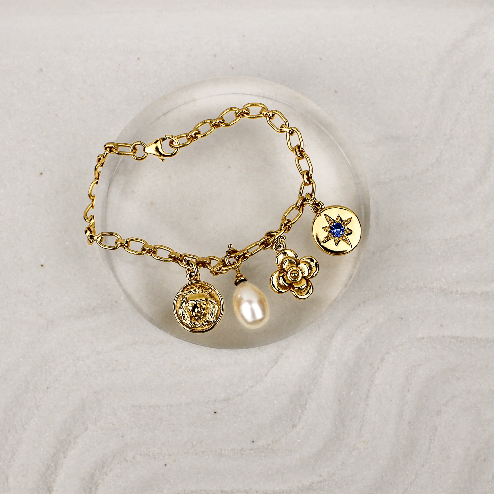 Archivist Bracelet 18k gold vermeil on clear dish over sand, multi-charm bracelet with lion, pearl, flower and north star charms, Ether Jewelry