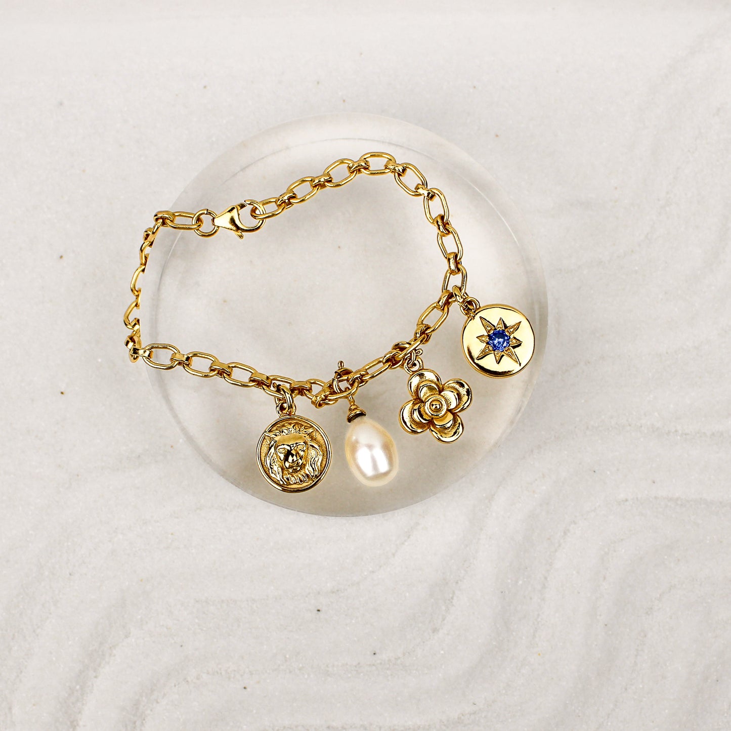 Archivist Bracelet 18k gold vermeil on clear dish over sand, multi-charm bracelet with lion, pearl, flower and north star charms, Ether Jewelry