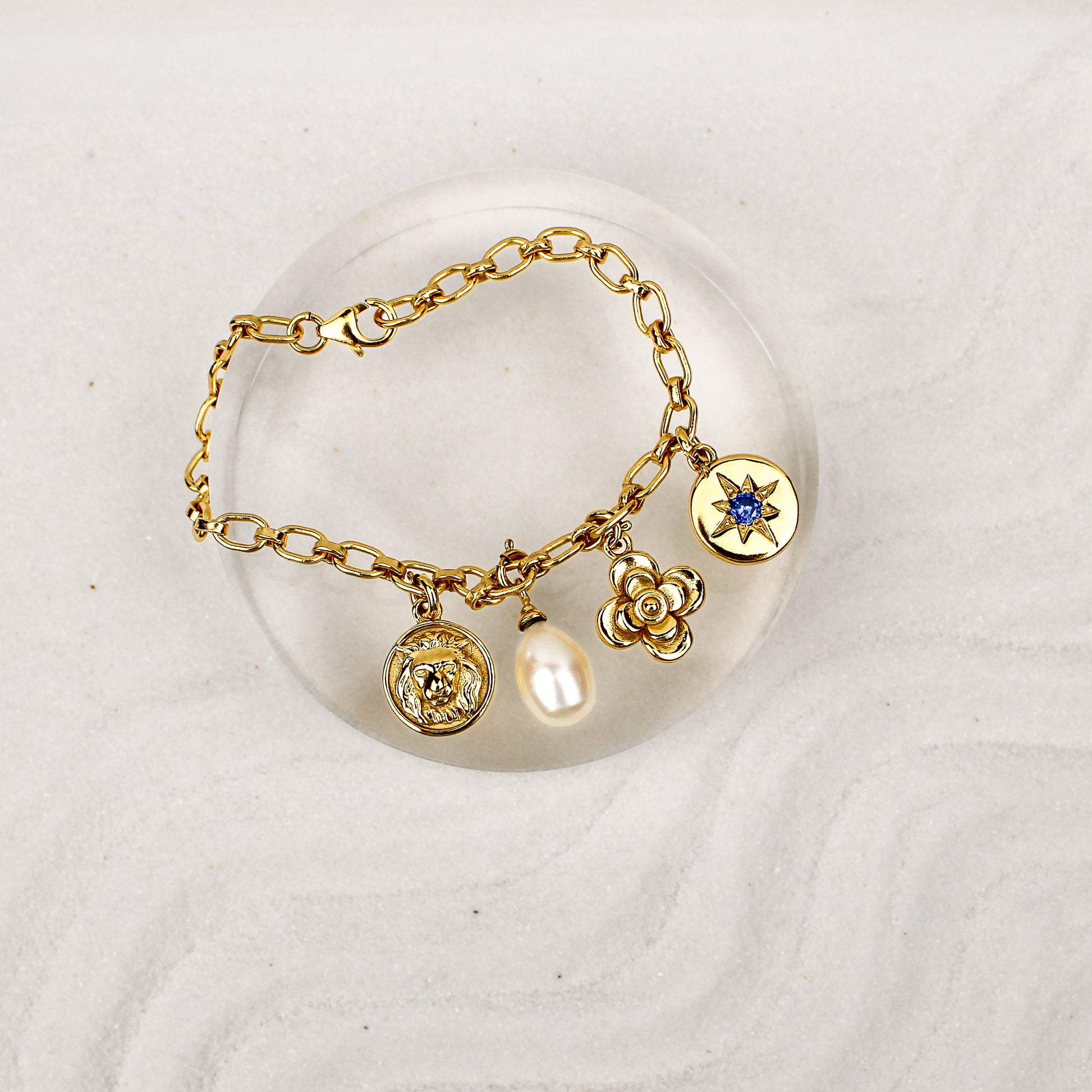 Archivist Bracelet 18k gold vermeil on clear dish over sand, multi-charm bracelet with lion, pearl, flower and north star charms, Ether Jewelry