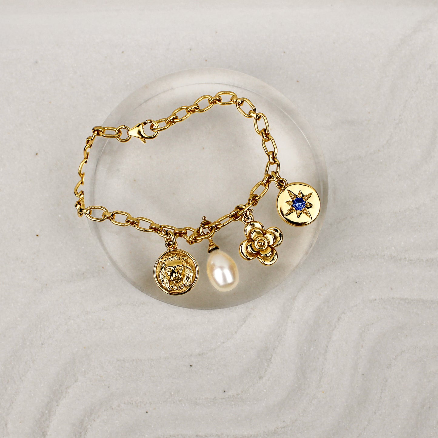 Gold archivist bracelet with charms on a white sand background and clear dish