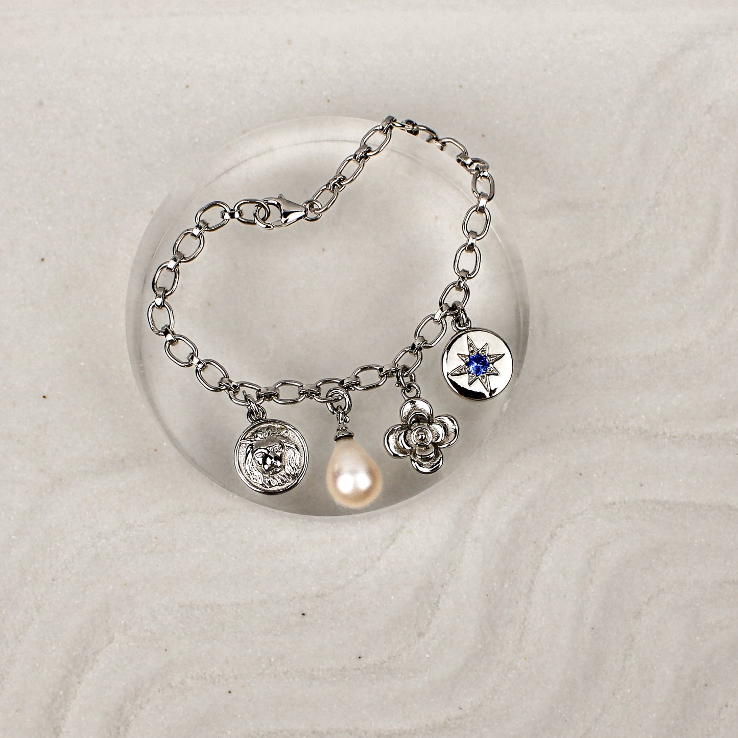 Archivist Bracelet sterling silver on clear dish over sand, multi-charm bracelet with lion, pearl, flower and north star charms, Ether Jewelry