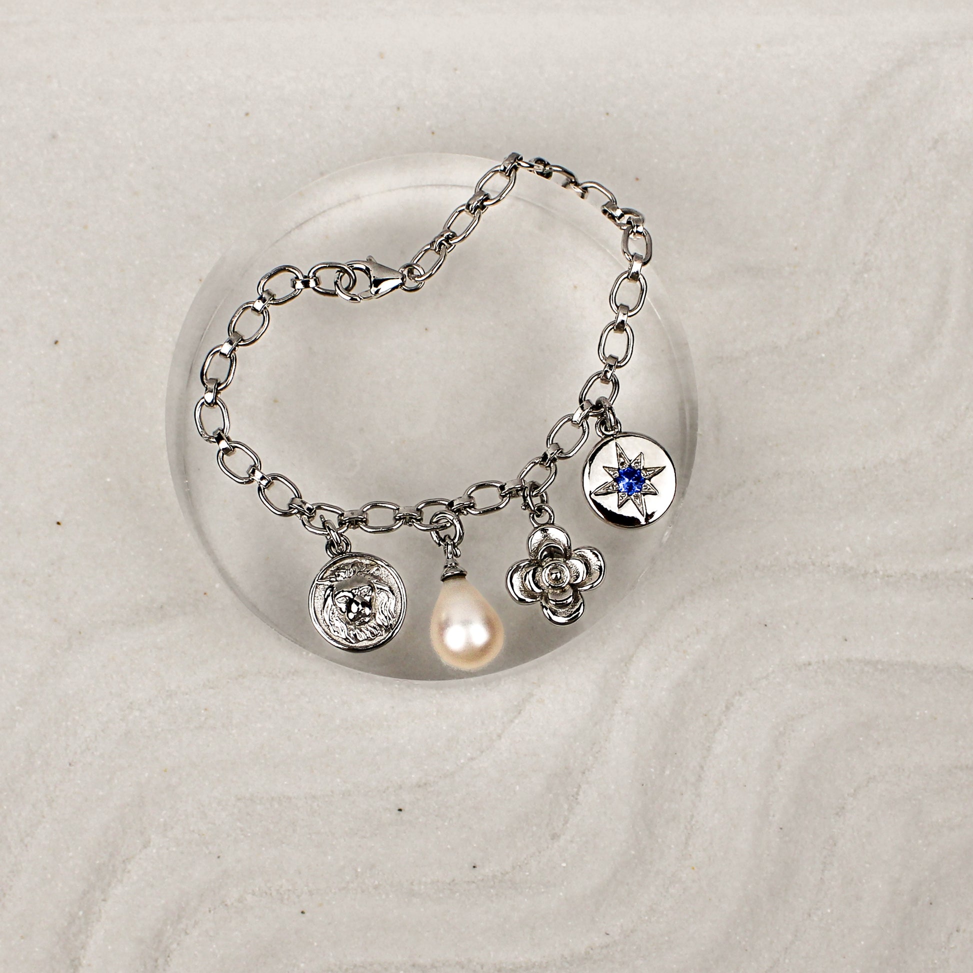 Archivist Bracelet sterling silver on clear dish over sand, multi-charm bracelet with lion, pearl, flower and north star charms, Ether Jewelry