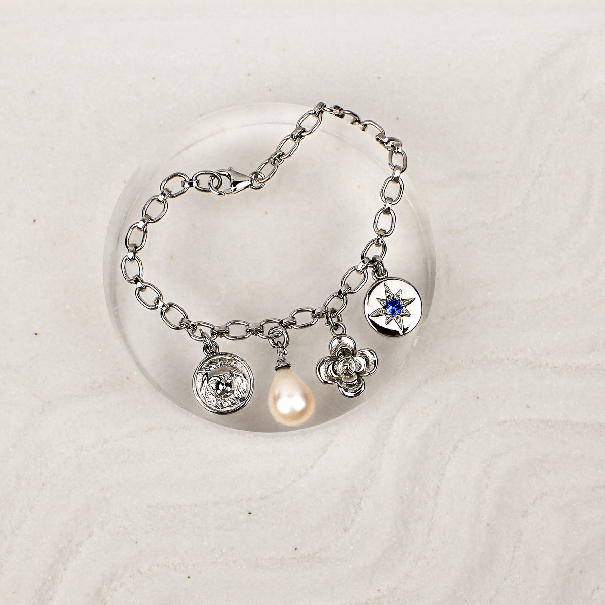 Archivist Bracelet sterling silver on clear dish over sand, multi-charm bracelet with lion, pearl, flower and north star charms, Ether Jewelry