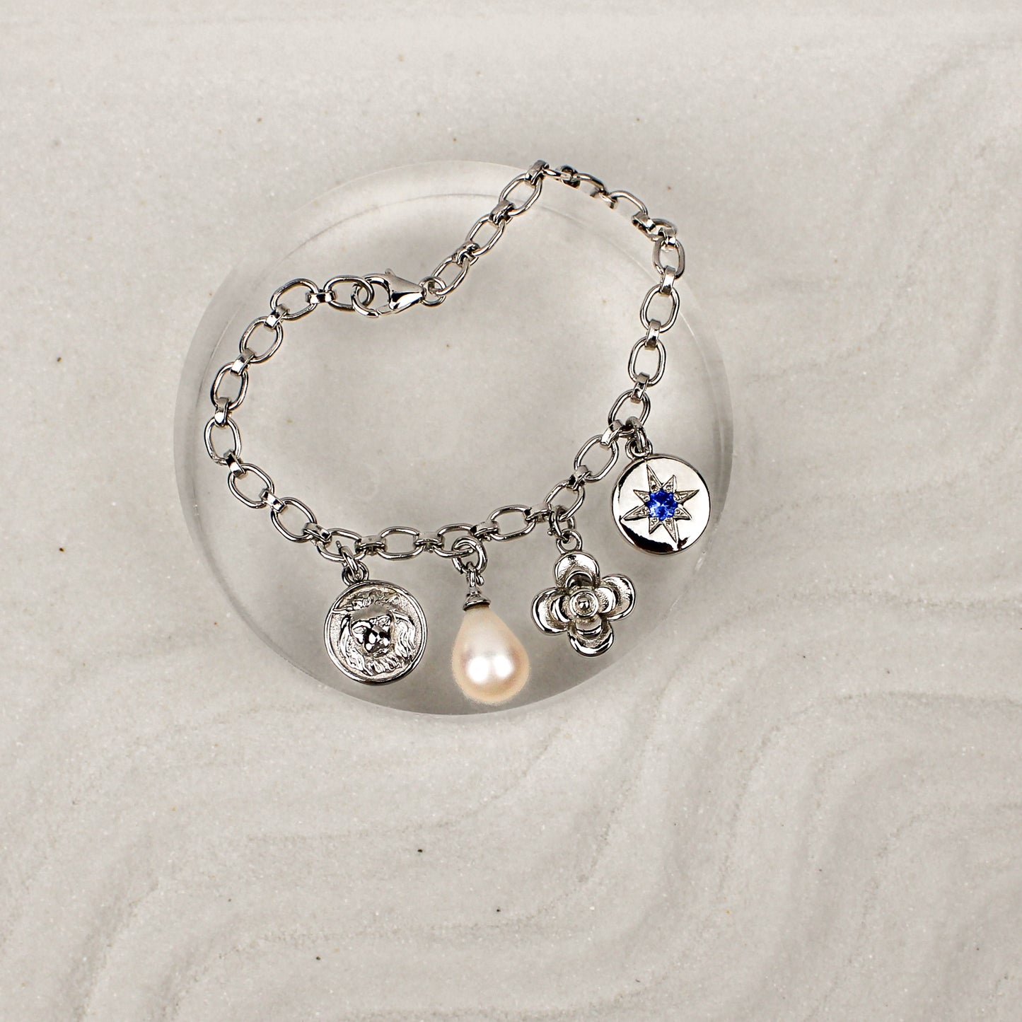 Silver bracelet with various charms on a white sand background and clear dish