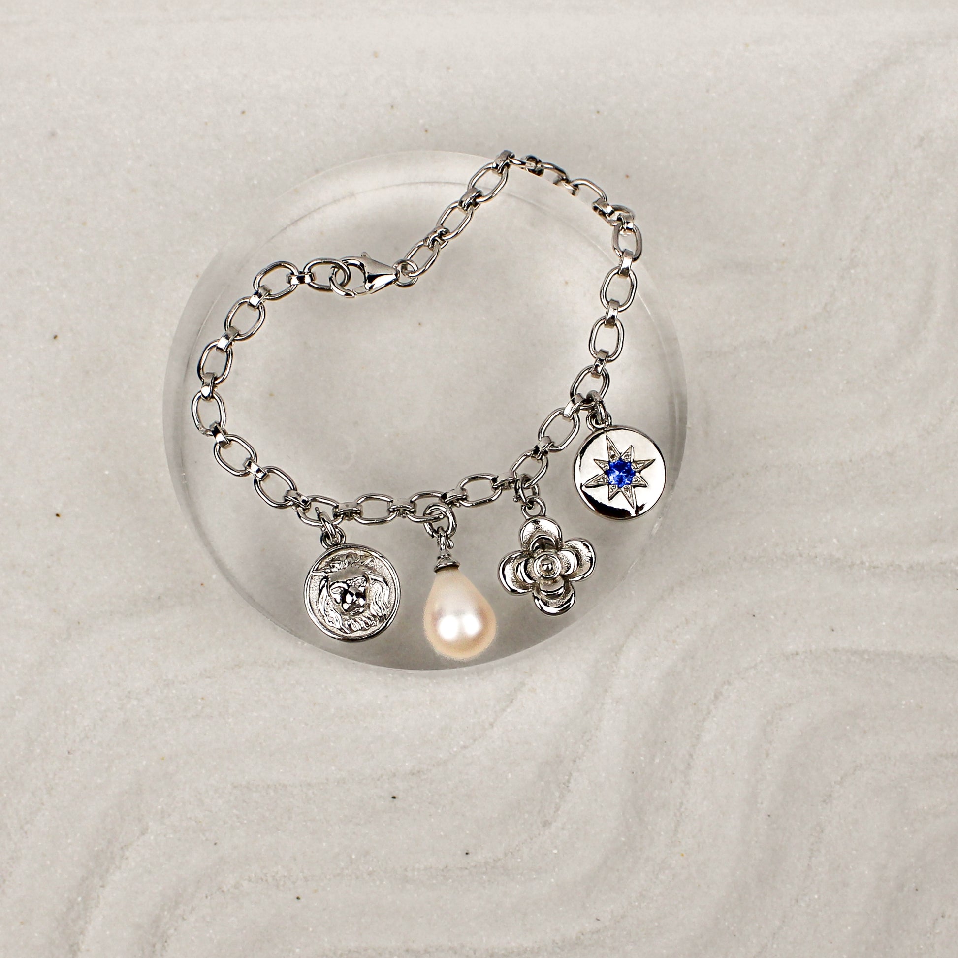 Silver bracelet with various charms on a white sand background and clear dish