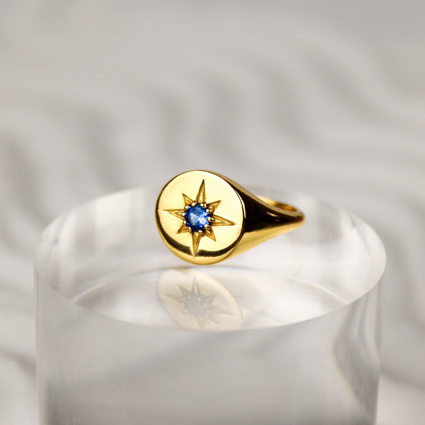Gold Wayfinder signet ring with a blue gemstone and star engraving on clear pedestal on a white sand background 