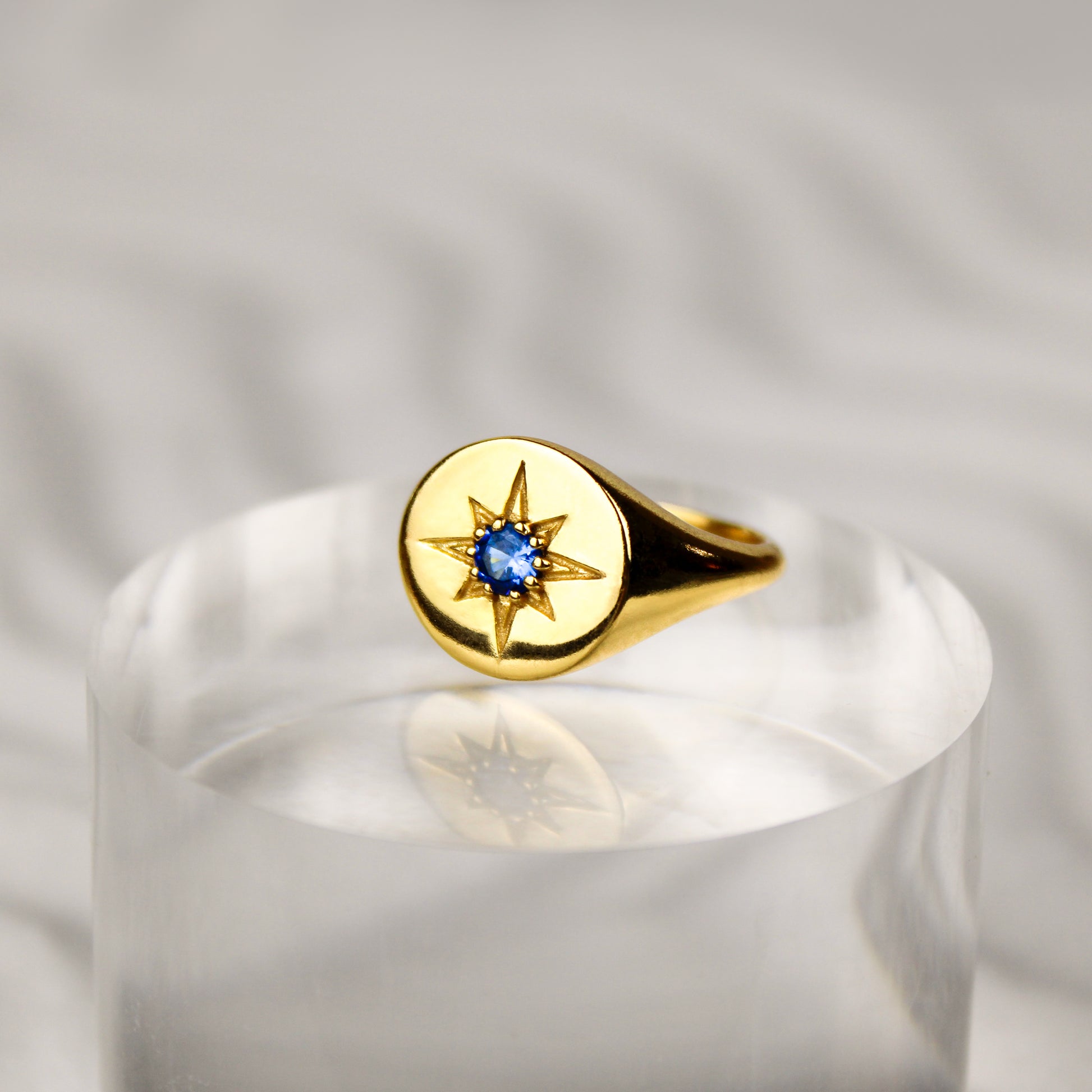 Gold Wayfinder signet ring with a blue gemstone and star engraving on clear pedestal on a white sand background 