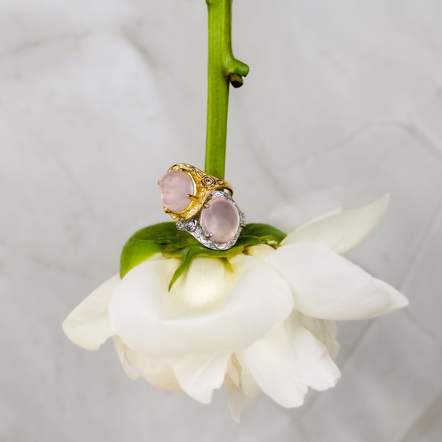 romantic heir signet Ring in gold and silver with pink quartz gemstone and flower engravings on a white flower stem against a light gray marble background