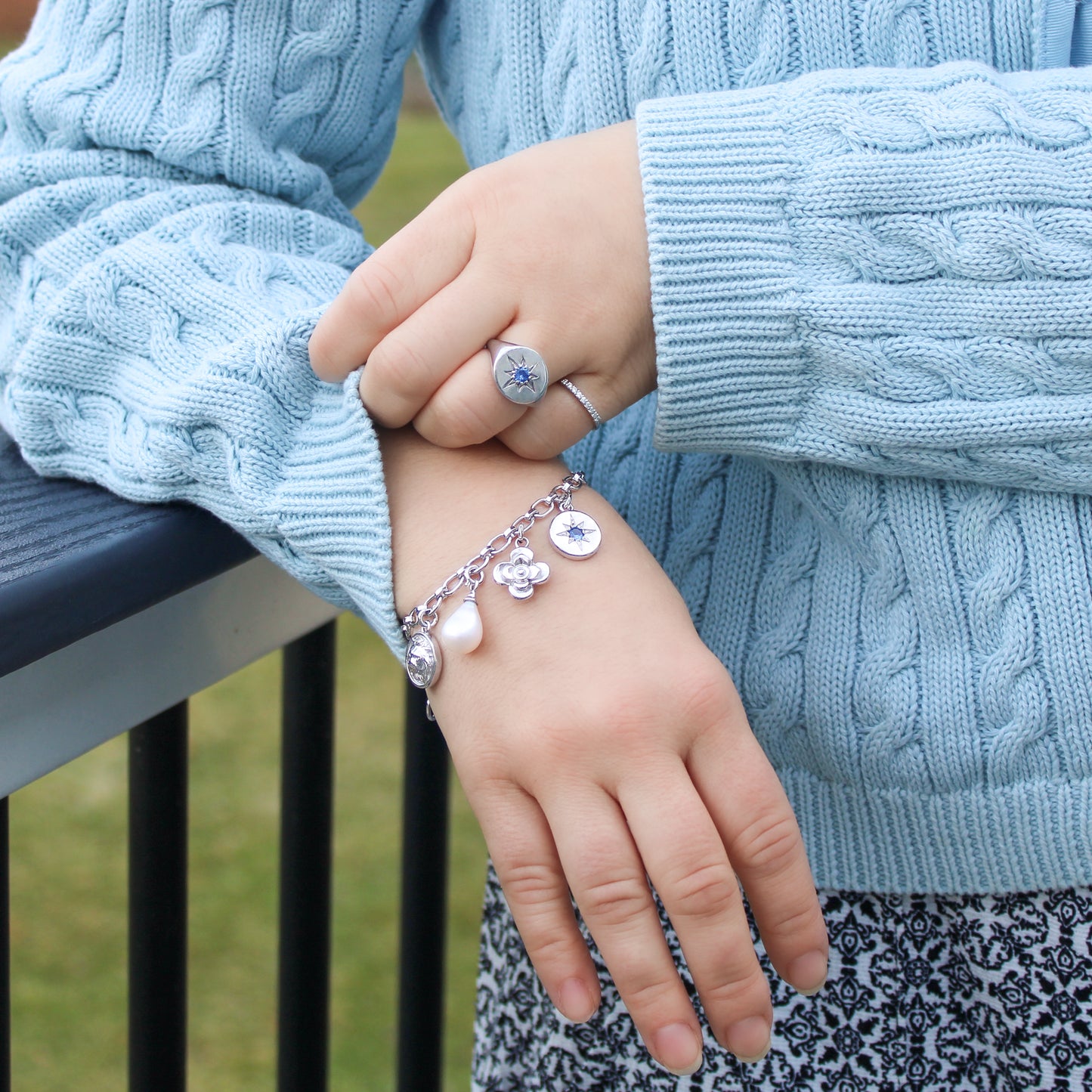 Close-up of a hand wearing a bracelet with charms and matching ring, holding another hand against a light blue sweater.