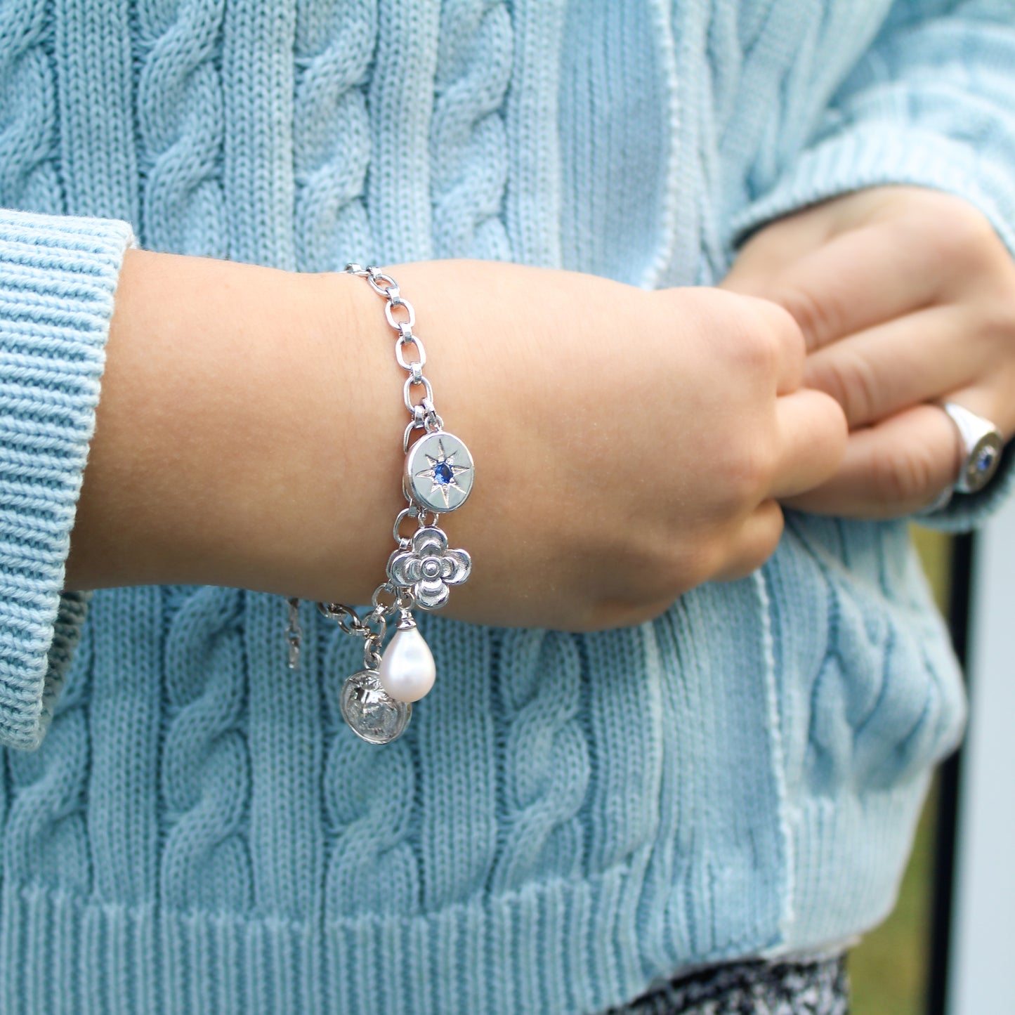 Close-up of a women's hand wearing a sterling silver archivist bracelet with charms against a light blue knitted sweater.