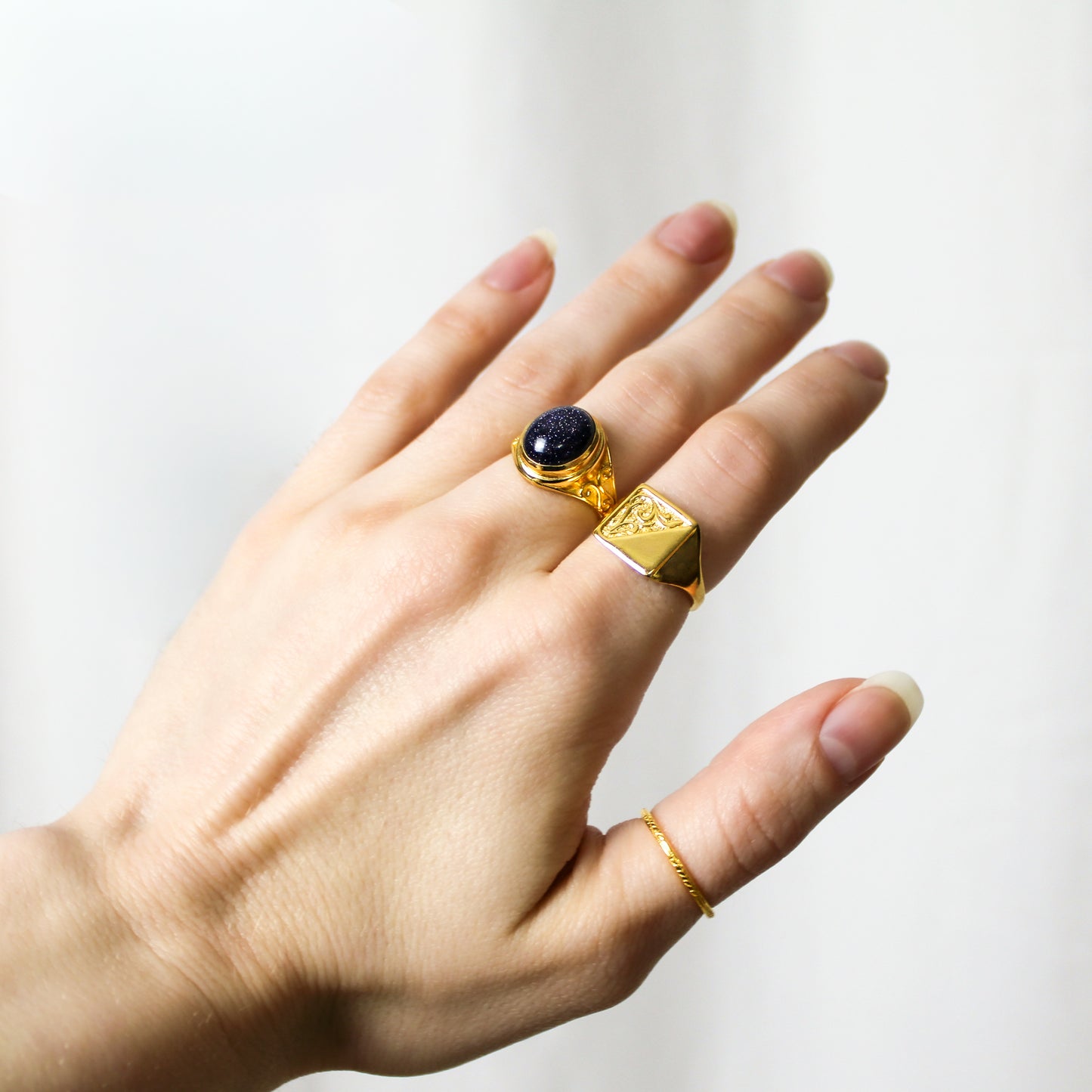 Hand wearing a gold ring with a dark blue sandstone gemstone, and all metal gold vermeil ring on a light background, Ether Jewelry