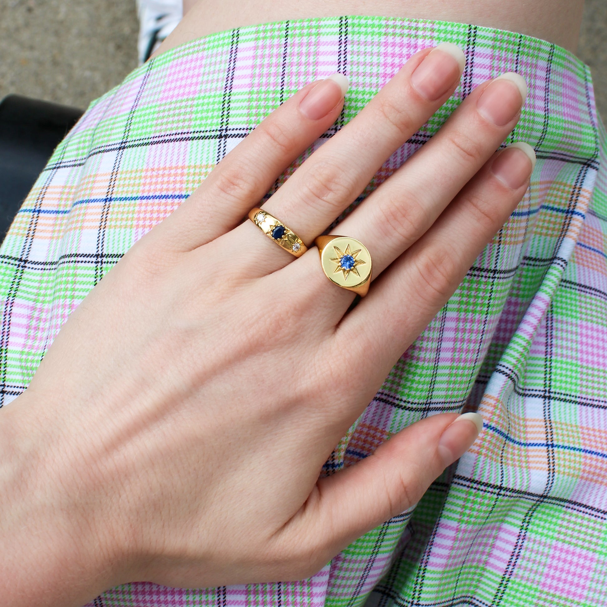 Top down view of hand wearing two gold rings with blue gemstones and starburst engraving against a plaid skirt, Ether Jewelry
