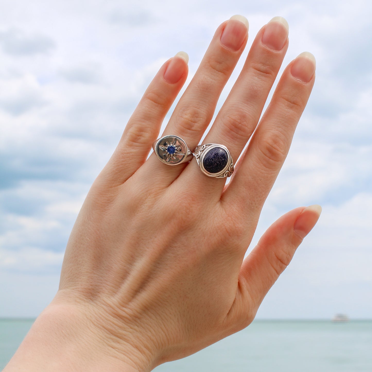 Hand wearing two sterling silver rings with blue sandstone and starburst engraving against a blurred beach background, Ether Jewelry
