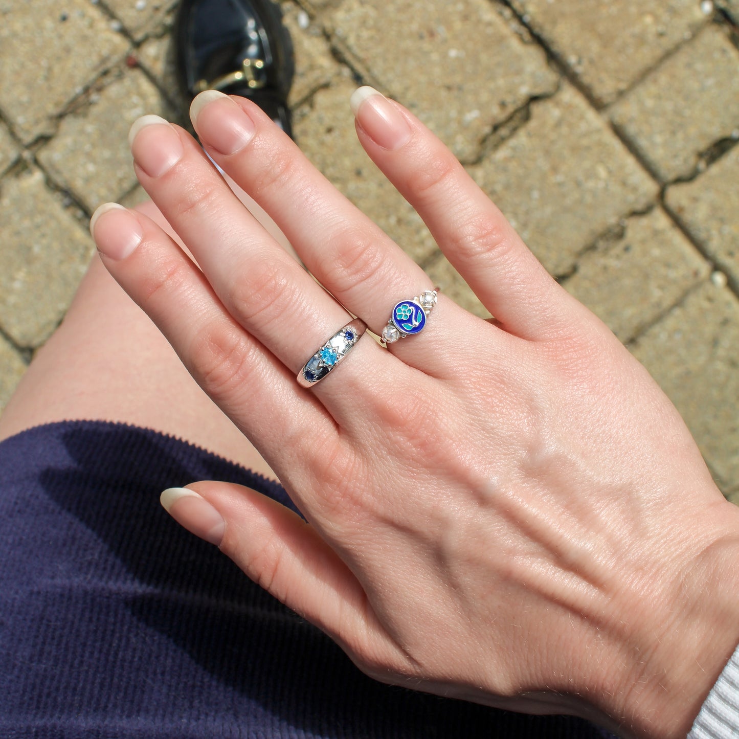 Close-up of a hand wearing two rings with blue gemstones and forget-me-not on a textured pavement background, Ether Jewelry