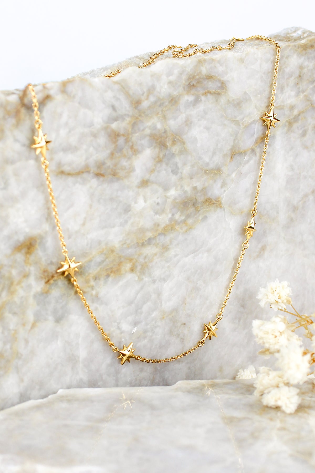 Juno Necklace in 18k gold vermeil, delicate cable chain with scattered solid north star charms, staged on marble with white flowers, Ether Jewelry