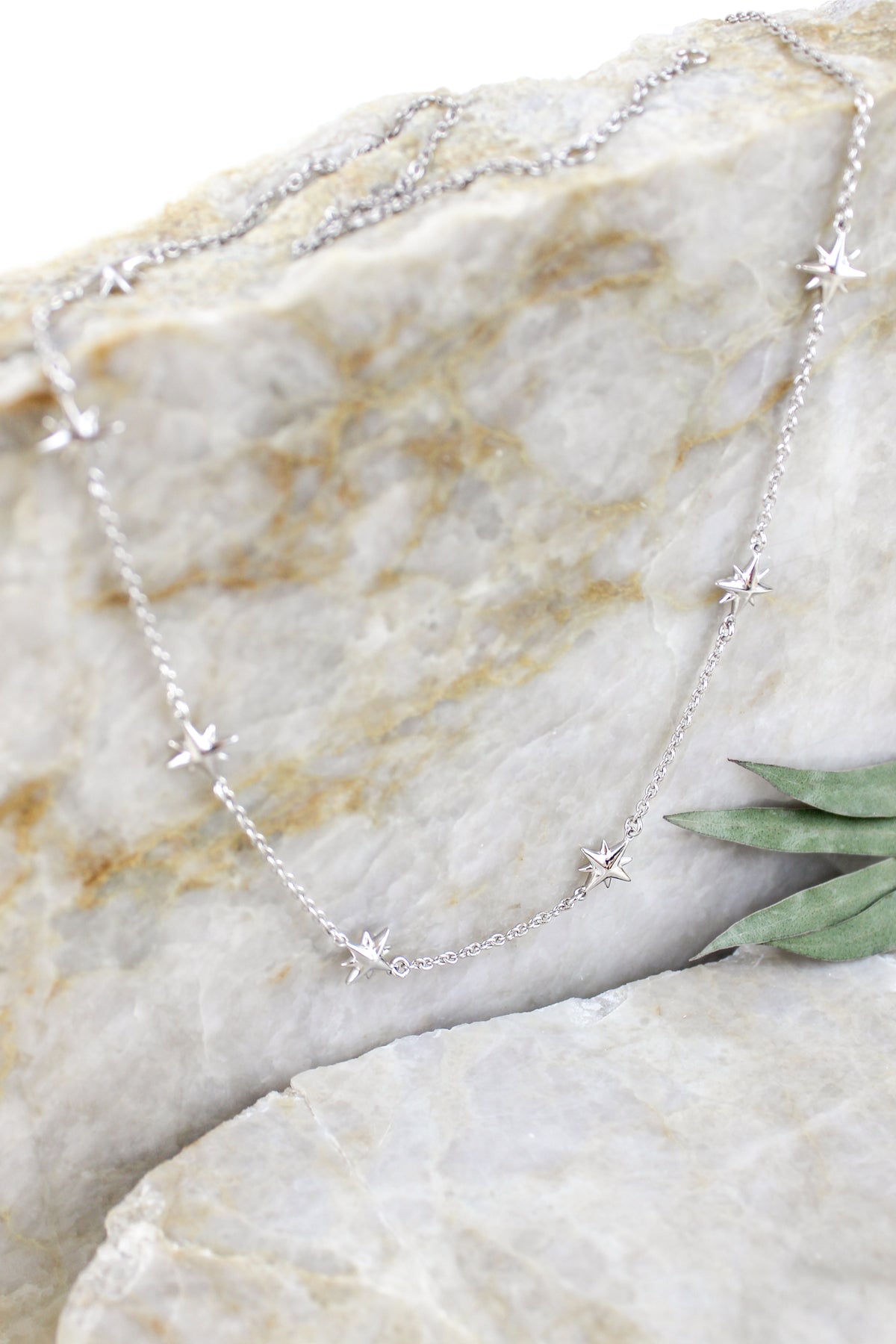 Juno Necklace in sterling silver, delicate cable chain with scattered solid north star charms, staged on marble with eucalyptus, Ether Jewelry