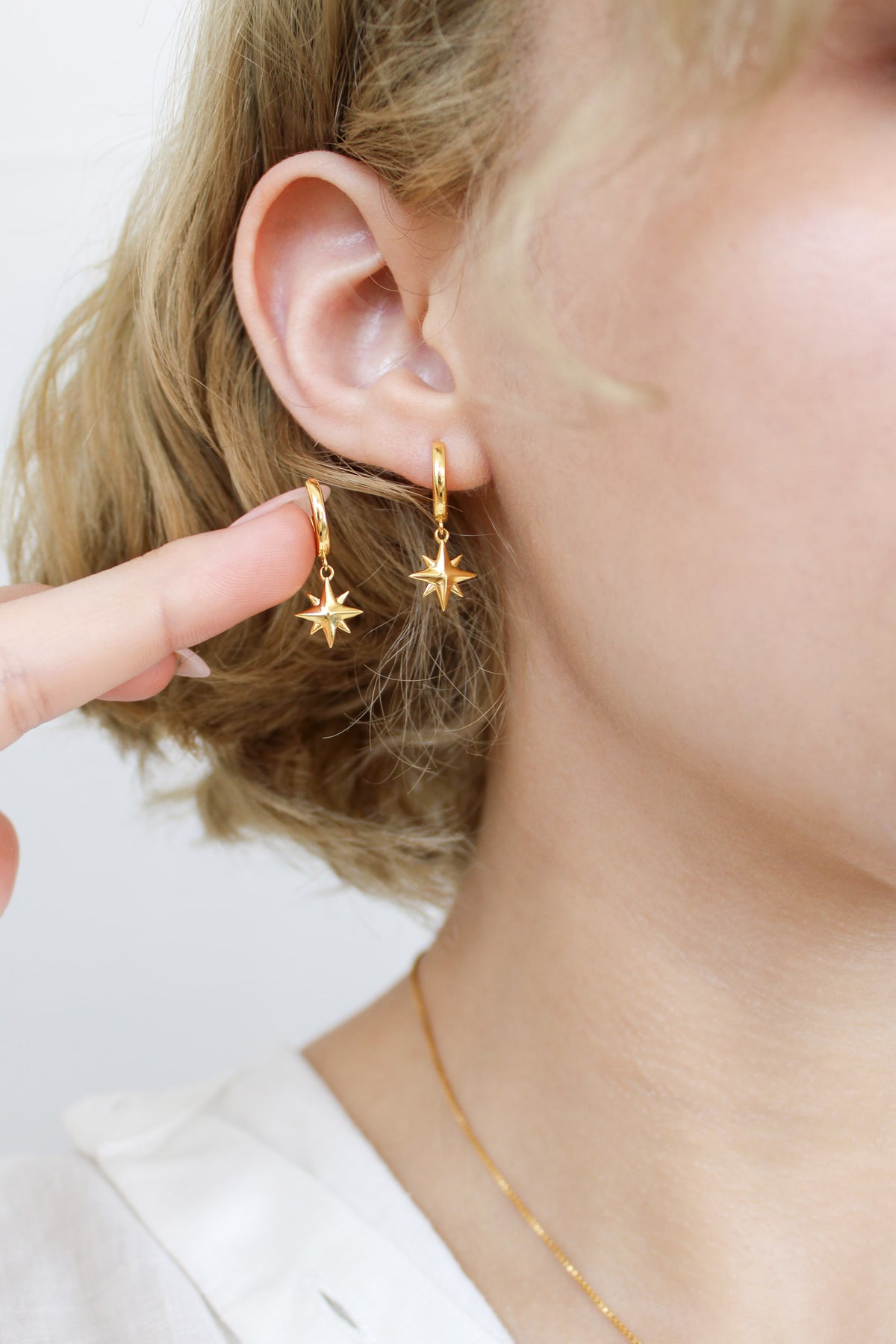 Stella Hoops 18k gold vermeil held on model fingertip next to earring in ear, north star charm drop huggie hoops, Ether Jewelry