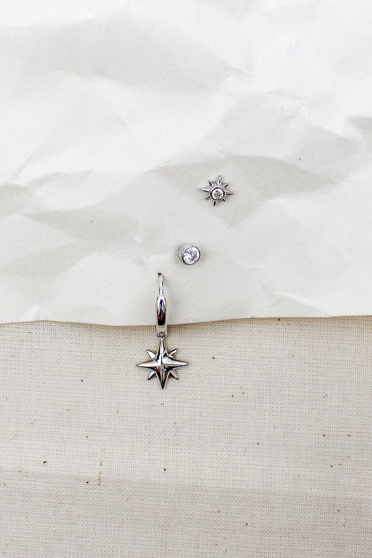 Stella Hoops sterling silver staged on paper and natural fabric with Vesta stud earrings and stella stud earrings, north star charm drop earrings, Ether Jewelry