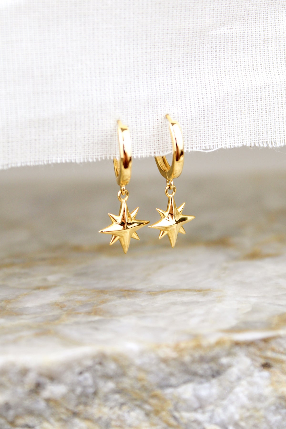 Stella Hoops 18k gold vermeil staged on white fabric above marble, north star charm drop huggie hoops, Ether Jewelry