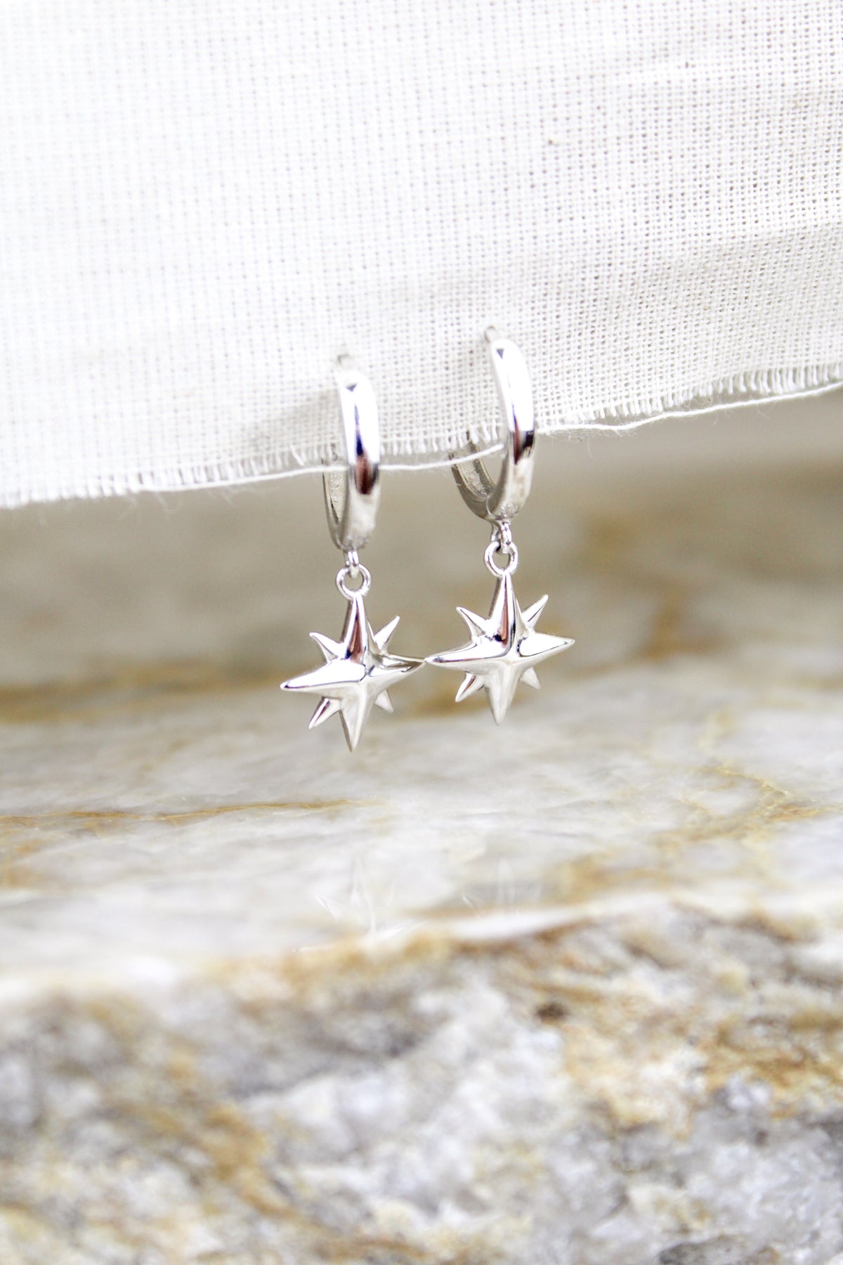 Stella Hoops sterling silver staged on white fabric above marble, north star charm drop huggie hoops, Ether Jewelry