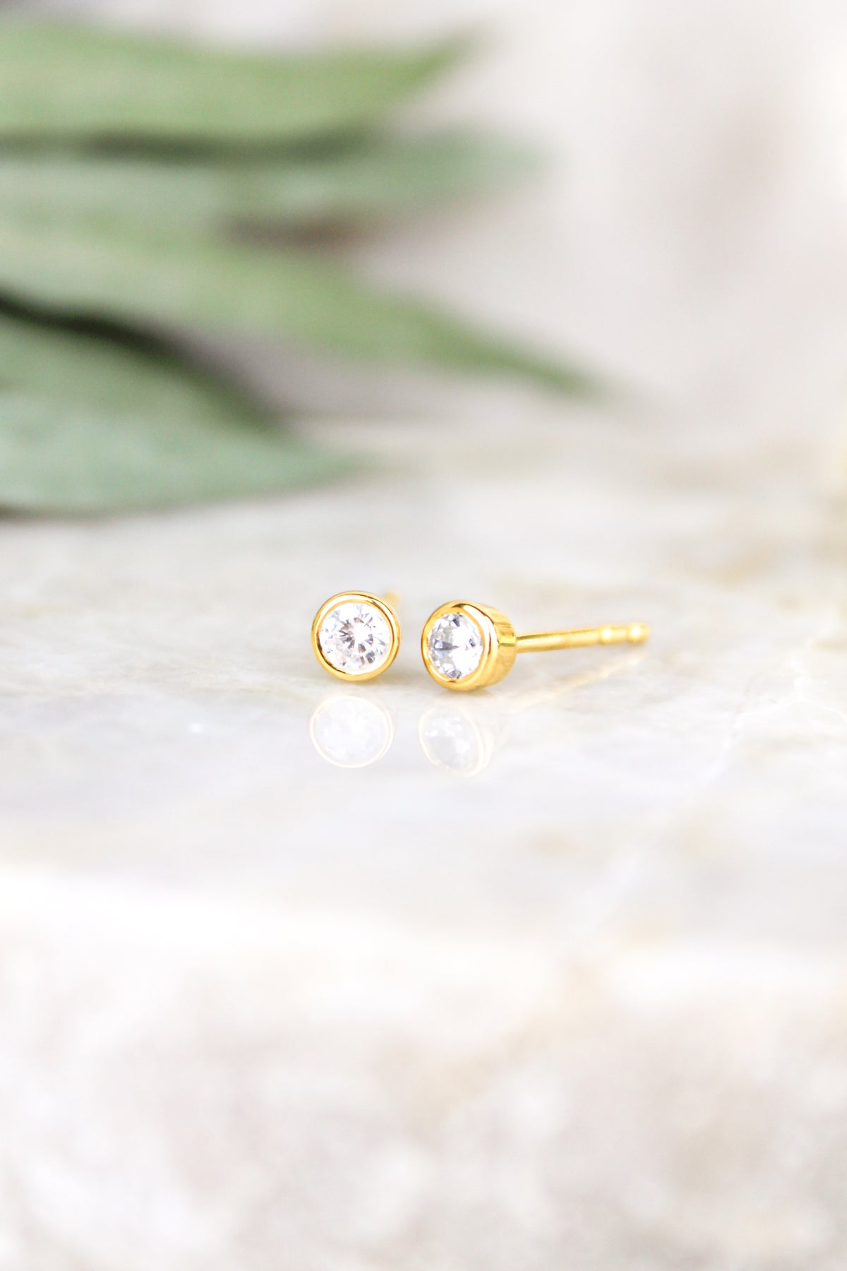 Vesta Stud Earrings 18k gold vermeil staged on marble with eucalyptus and white flowers, bezel-set CZ studs, Ether Jewelry
