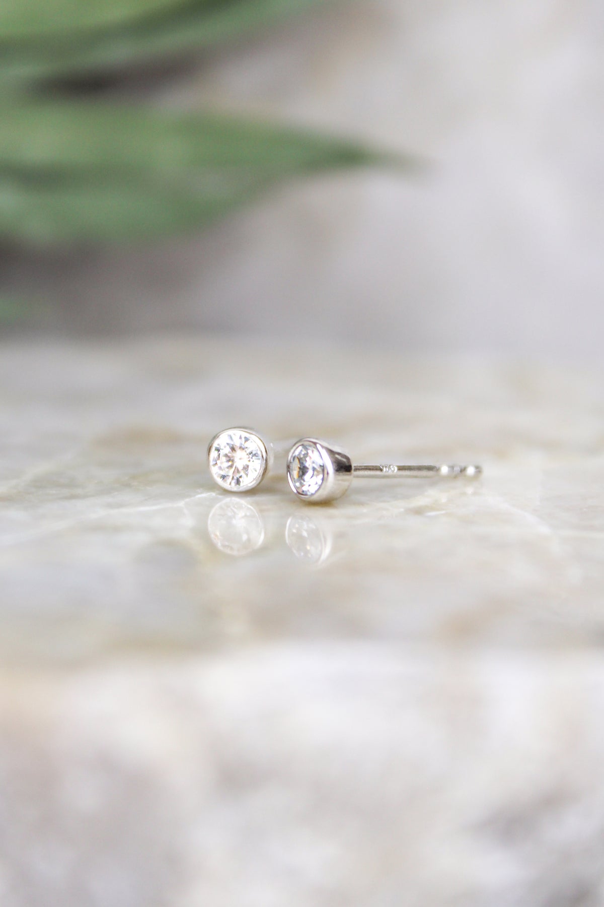 Vesta Stud Earrings sterling silver staged on marble with eucalyptus and white flowers, bezel-set CZ studs, Ether Jewelry