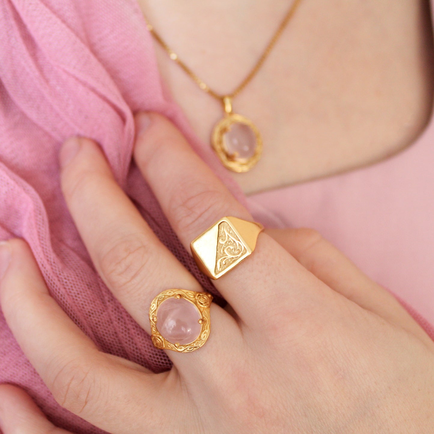Gold signet ring with pink quartz stone and flower engravings worn on a finger against a pink fabric background