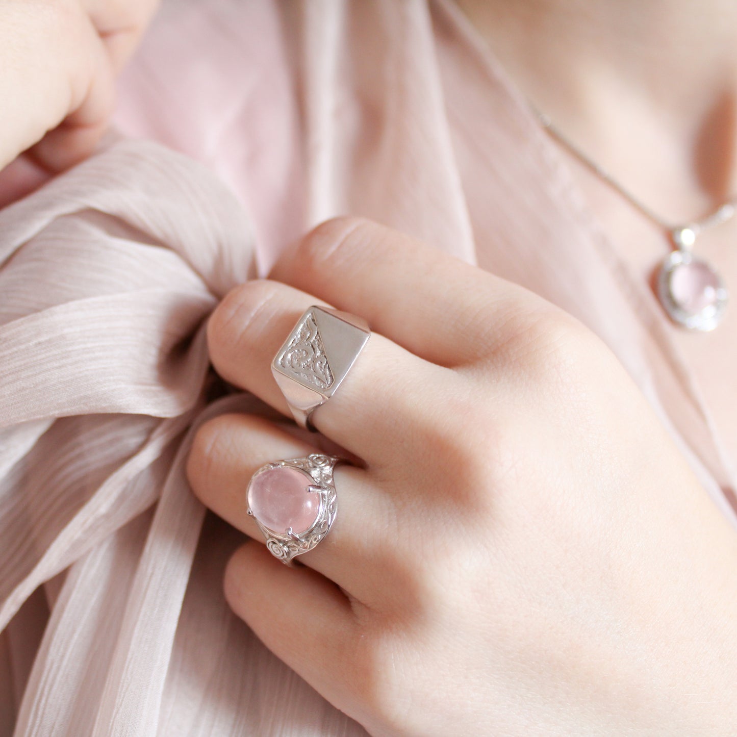 Close-up of a model's hand wearing two romantic heir signet rings with pink quartz and hand engravings on pink fabric