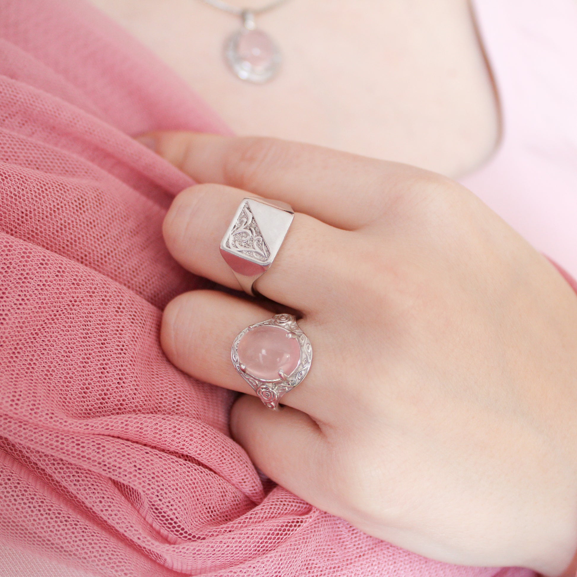 Model's hand wearing two romantic heir signet rings with hand engravings on pink fabric in the background