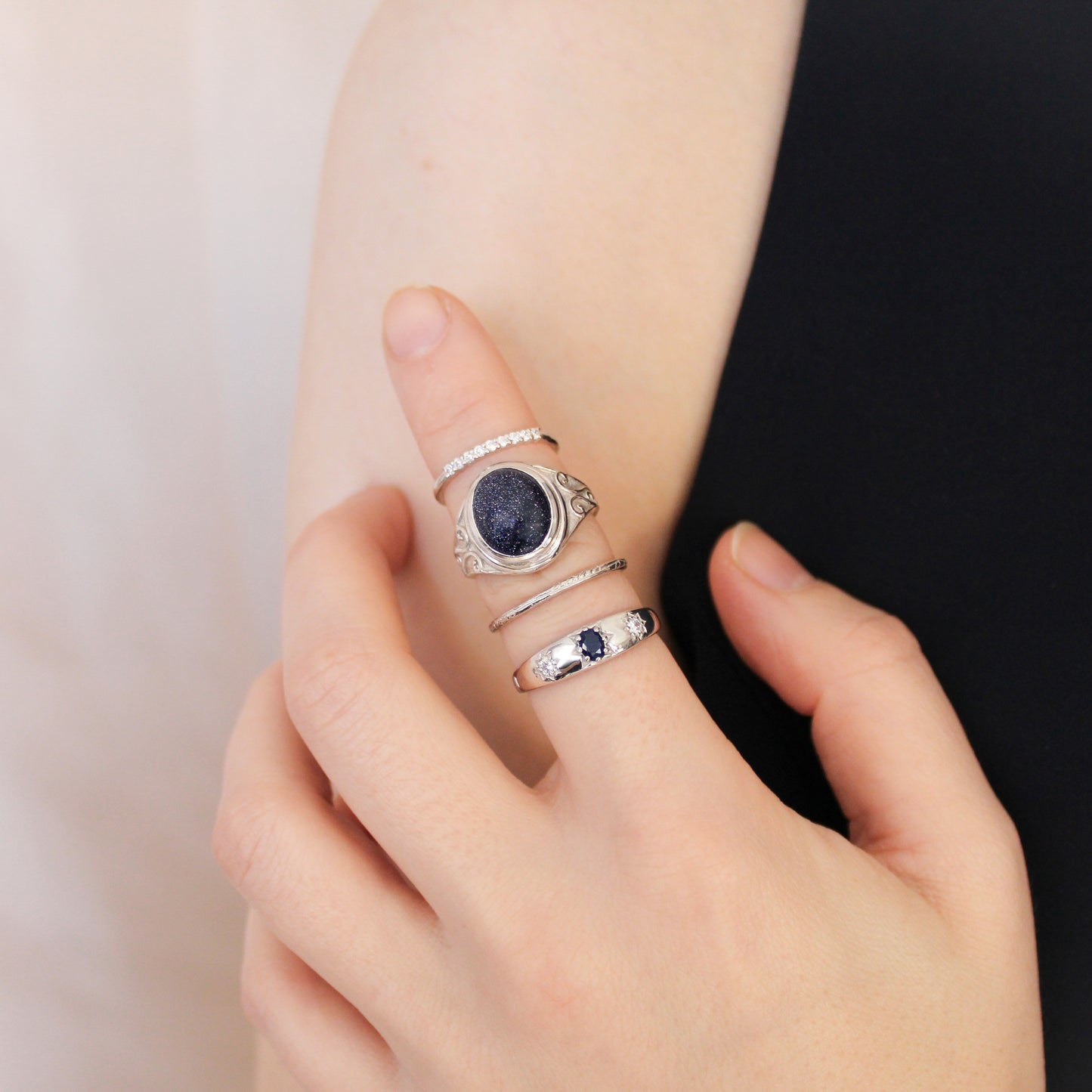 Hand wearing multiple sterling silver rings and night keeper signet ring with a dark blue sandstone on a neutral background