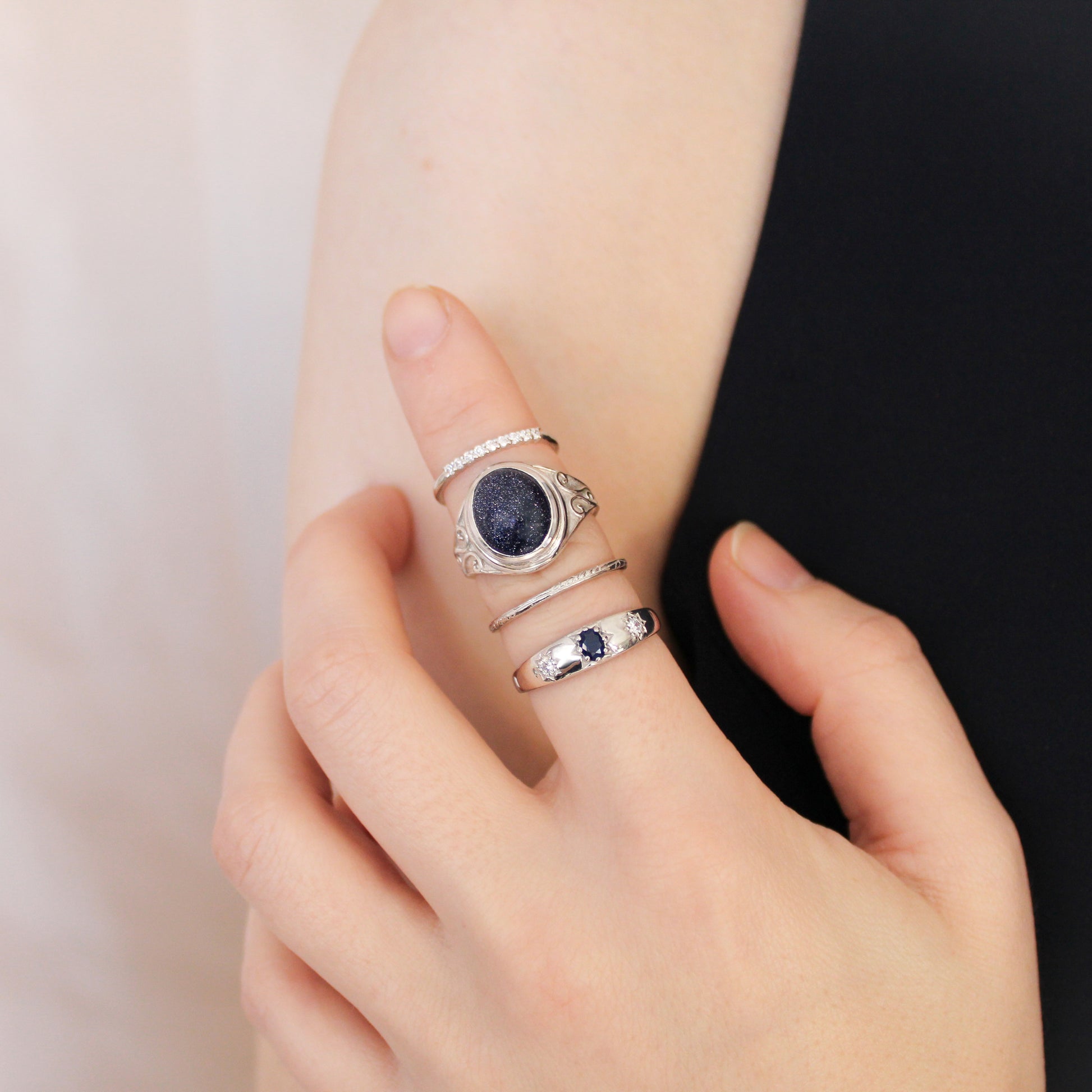 Hand wearing multiple sterling silver rings and night keeper signet ring with a dark blue sandstone on a neutral background