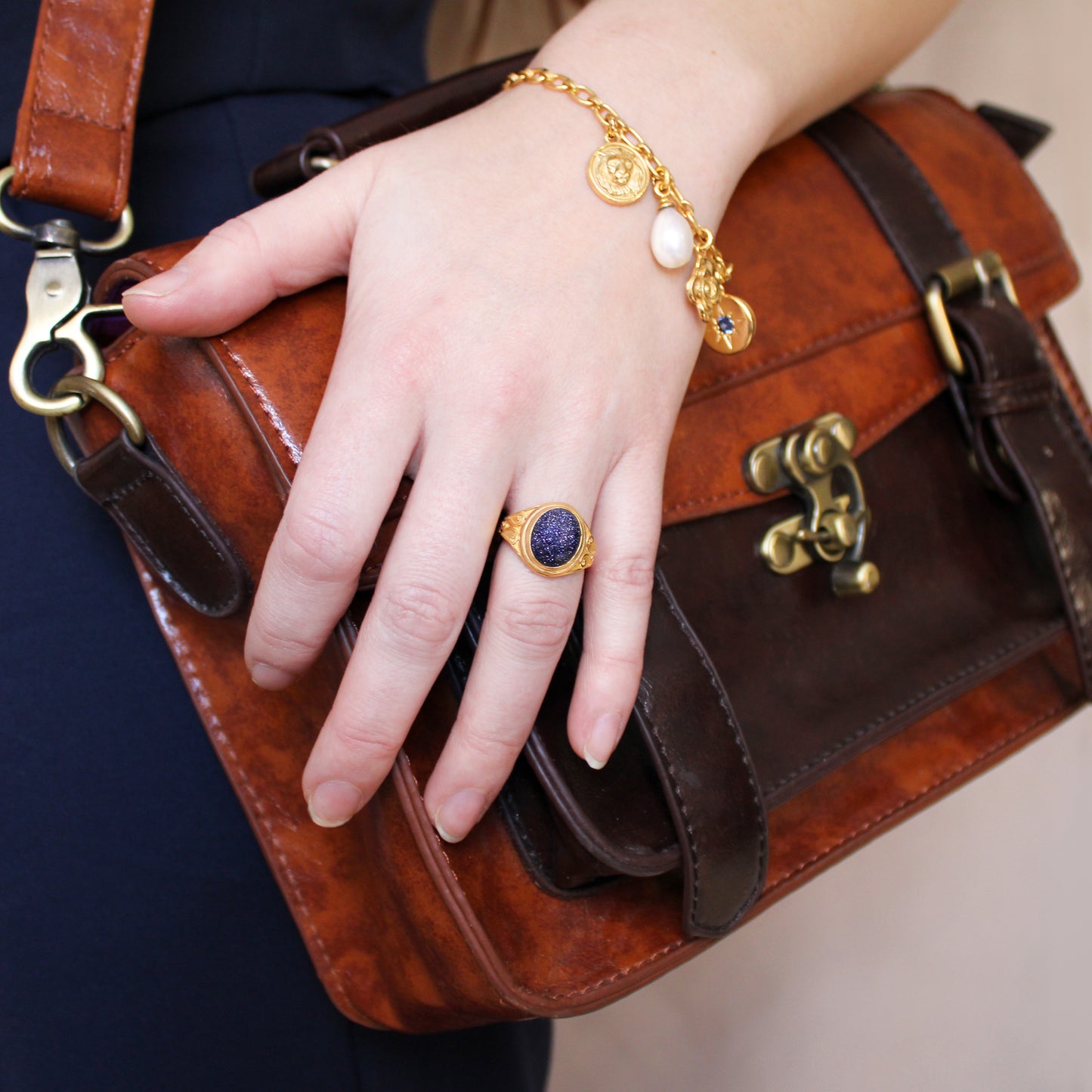 model's Hand holding a brown leather bag with a gold bracelet and night keeper signet ring with blue sandstone gemstone