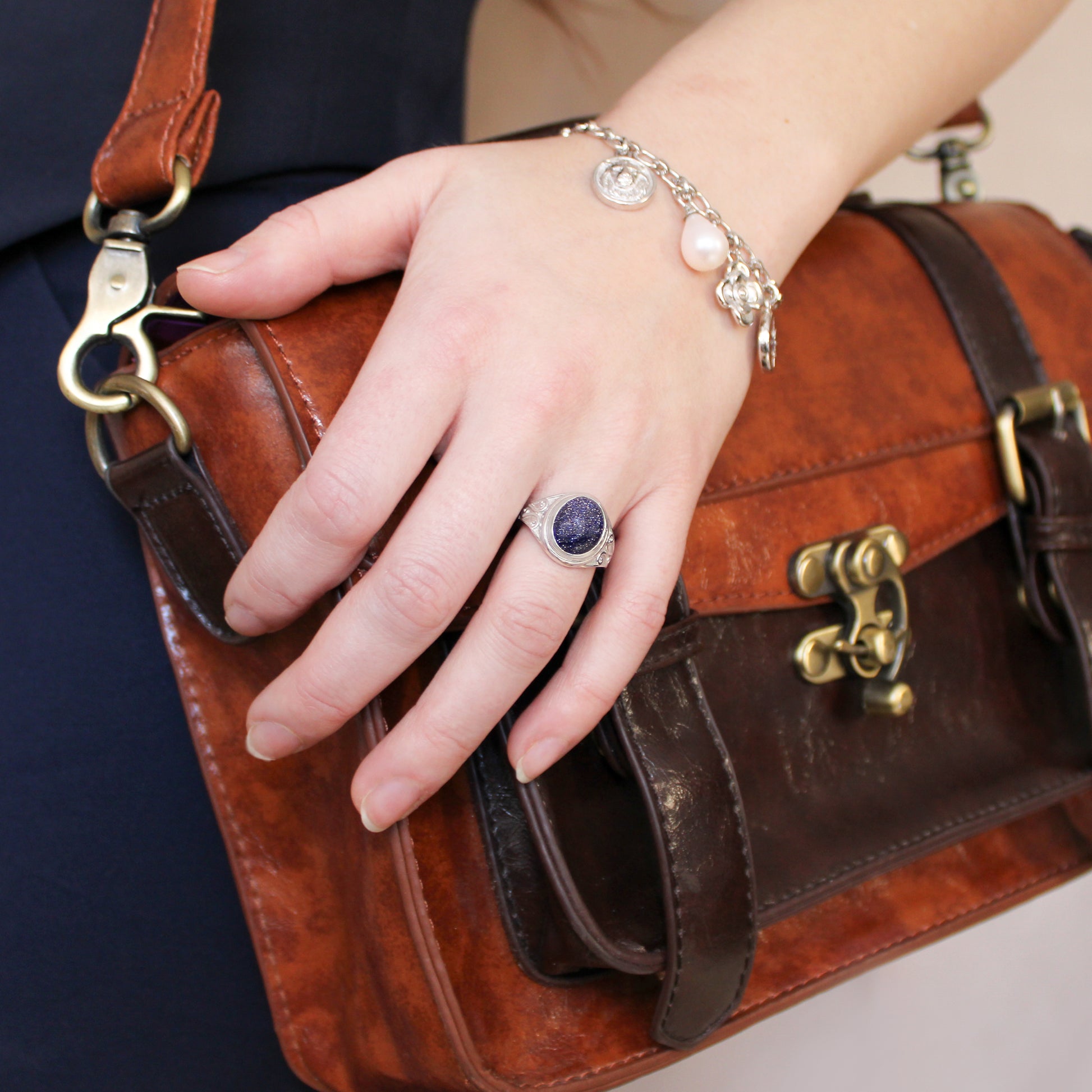 Hand holding a brown leather handbag with a close-up of a silver night keeper signet ring and charm bracelet.