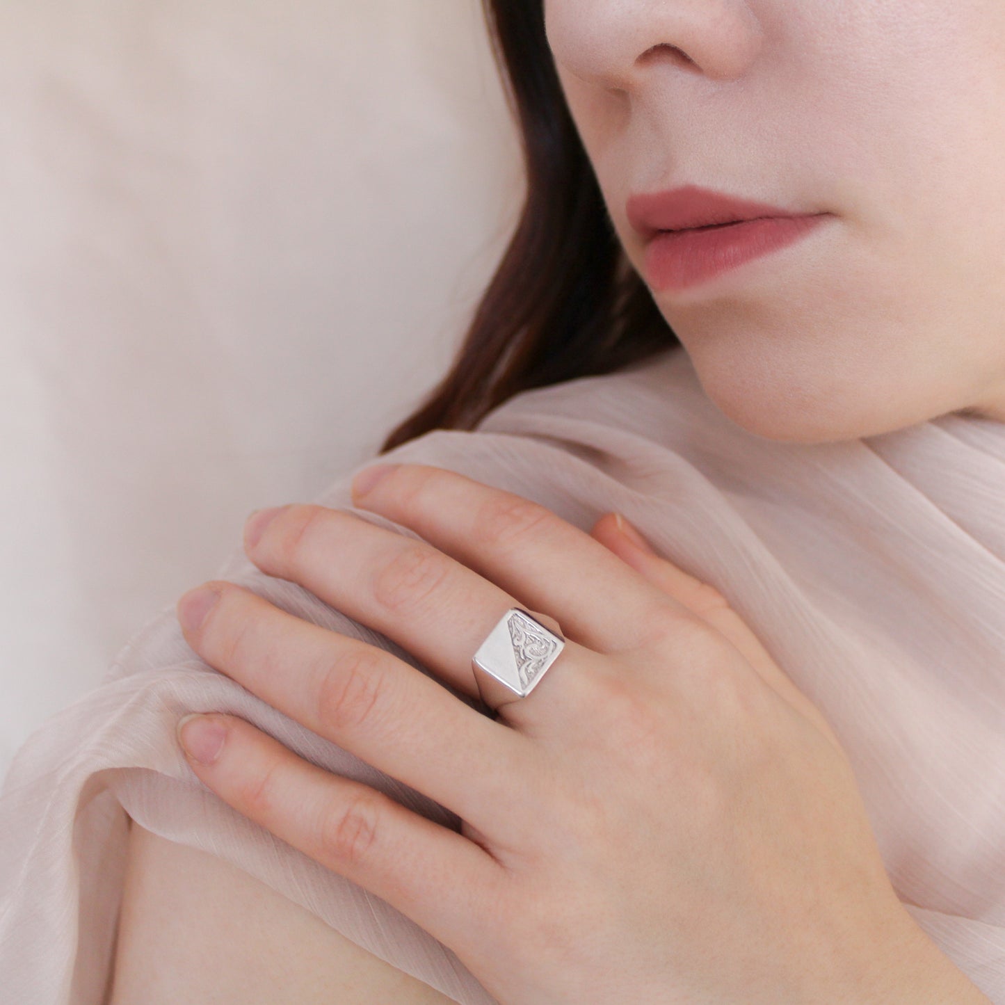 Close-up of a hand wearing a sterling silver architect signet ring with a intricate design, held against a soft pink background.