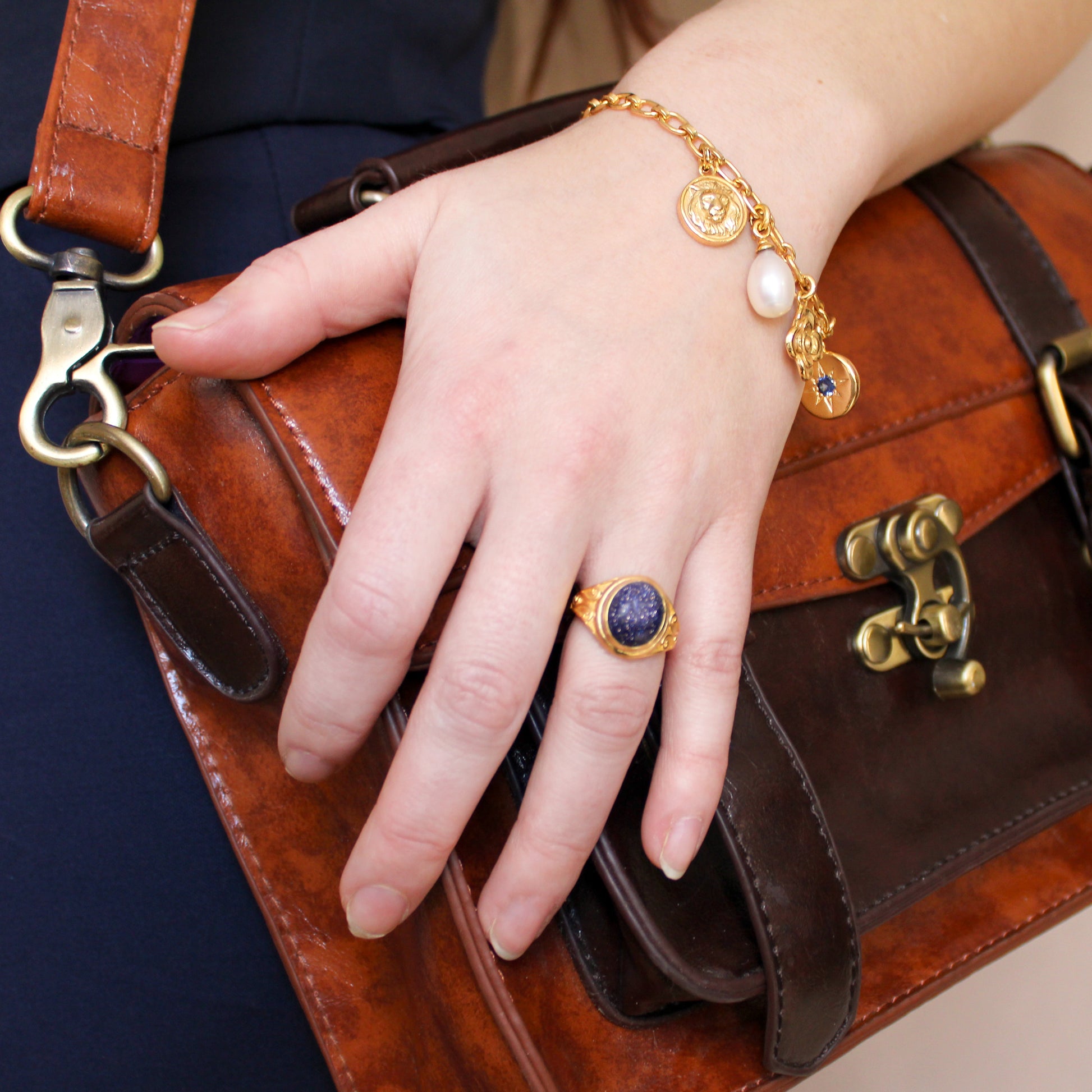 Hand with gold night keeper ring and archivist bracelet holding a brown leather bag