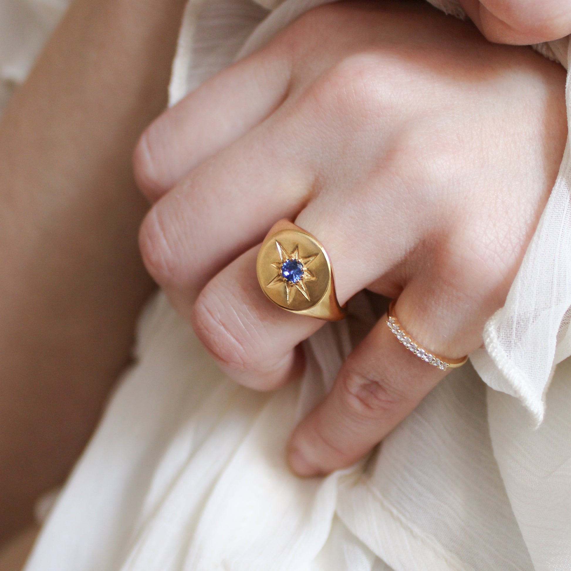 Close-up of a hand wearing a gold Wayfinder signet ring with a blue gemstone and star engraving on a white fabric background